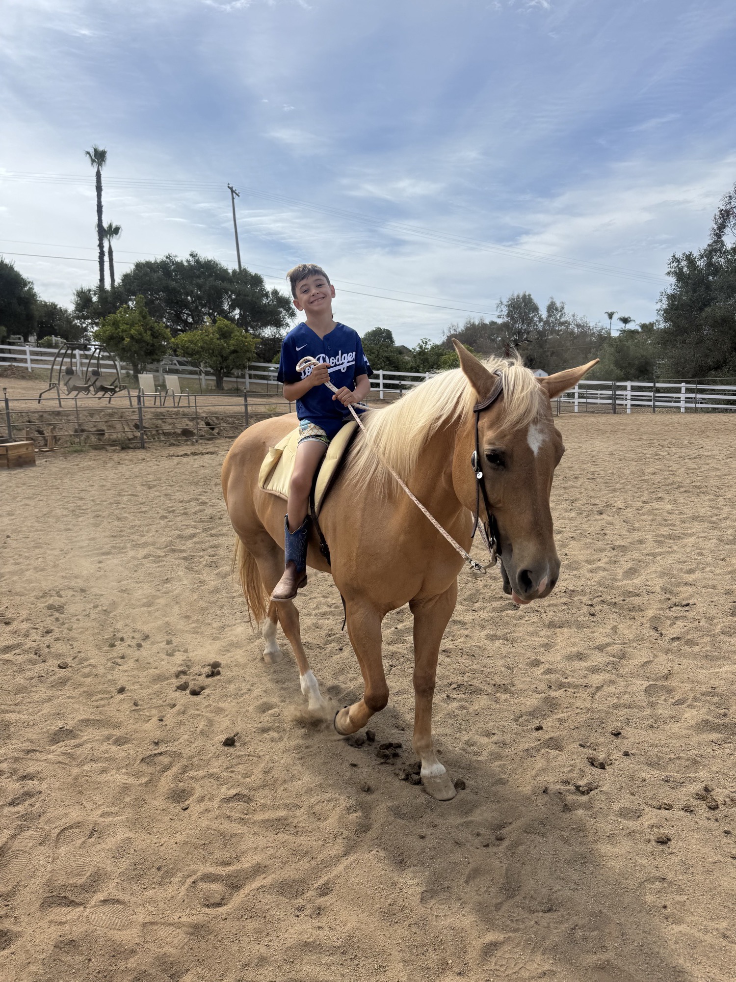 A smiling boy in a blue Dodgers shirt sits bareback on a palomino horse in the sandy arena